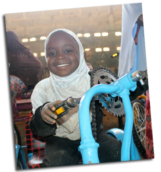 A young girl is sitting on her wheelchair, holding hand pedals made with bicycle parts.